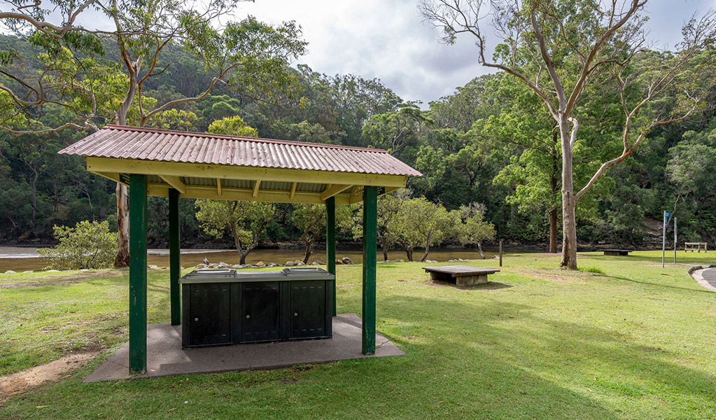 Sheltered barbecues next to a picnic platform on a lawn at Apple Tree picnic area with a creek and Ku-ring-gai Chase National Park bushland in the background. Credit: John Spencer &copy; DCCEEW