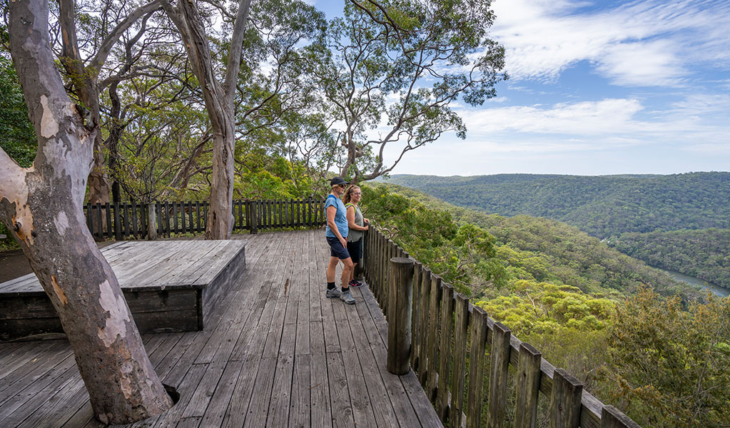Two people stand at a timber fence atop Kalkari lookout timber platform, overlooking Cowan Creek and a forested valley in Ku-ring-gai Chase National Park. Photo: Johny Spencer &copy; DCCEEW