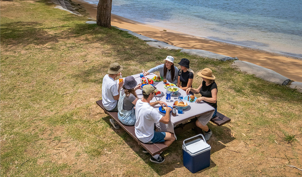 A group of people eating at a picnic table by the water at The Basin campground. Credit: John Spencer/DCCEEW  &copy; DCCEEW