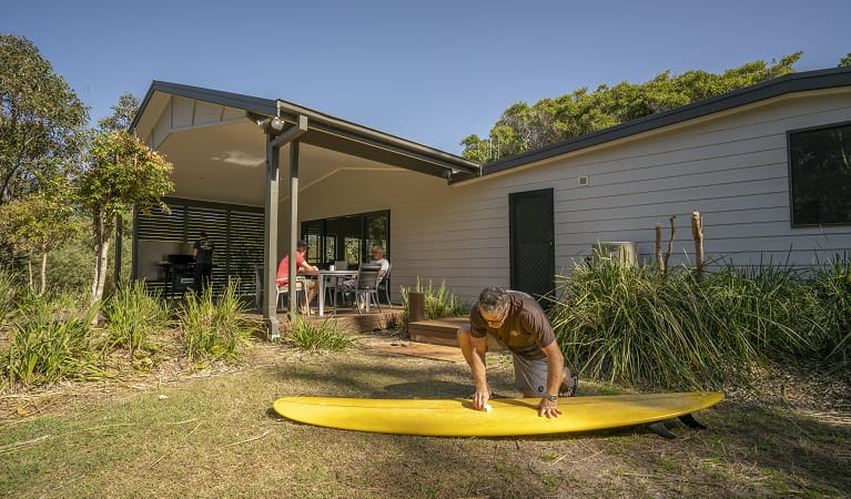 A man cleaning his surfboard on the grass nearby by the outdoor dining area at Plomer Beach House, Limeburners Creek National Park. Photo: John Spencer/OEH