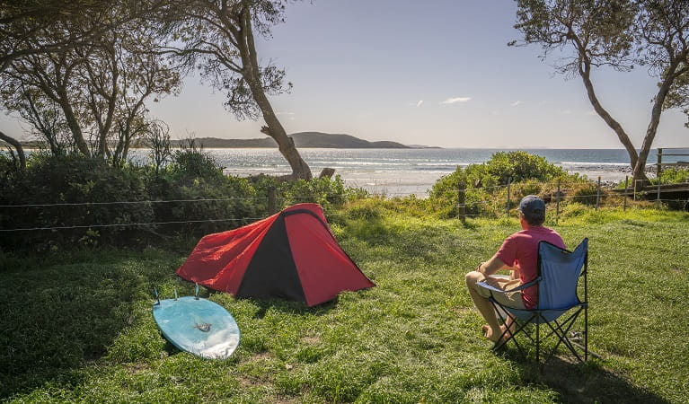 Man sitting beside his tent looking out at the ocean, at Limeburners Creek National Park. Photo: John Spencer/OEH