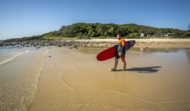 Man holding his surf board walking along the beach at Point Plomer campground in Limeburners Creek National Park. Photo: John Spencer/OEH