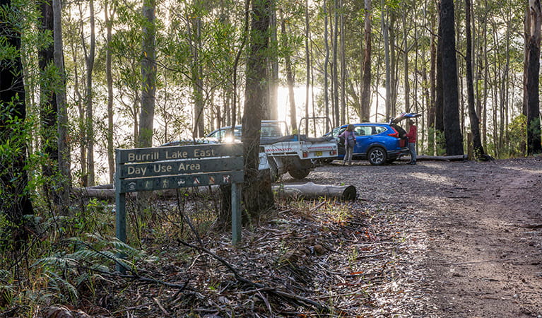 Sunlight reflects off the lake surface as 2 people unload their car at the parking area for Burrill Lake East picnic area. Photo: Michael Van Ewijk/DPIE