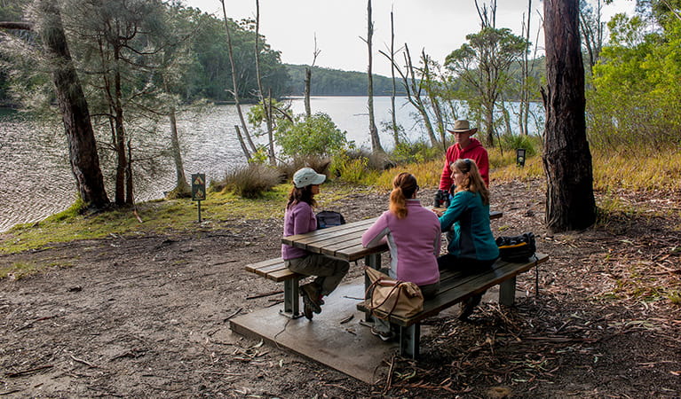 Group of 4 people at a picnic table in bushland setting next to Burrill Lake. Photo: Michael Van Ewijk/DPIE