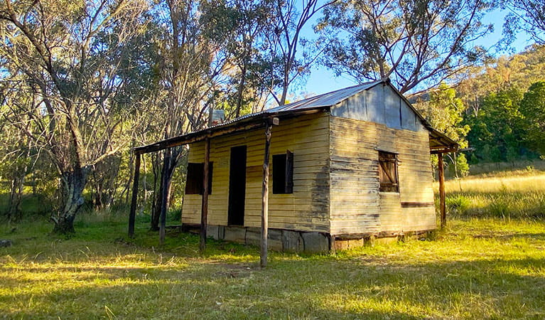 Side view of Scutts Hut set in grassy clearing with hills and bush land in the background. Photo credit: Lauren Sparrow &copy; DPIE