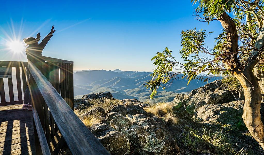 A visitor throws their hands in the air while standing on Governor lookout, with a vista of mountains, ridges and valleys into the distance. Photo Credit: Simone Cottrell  &copy; DPIE