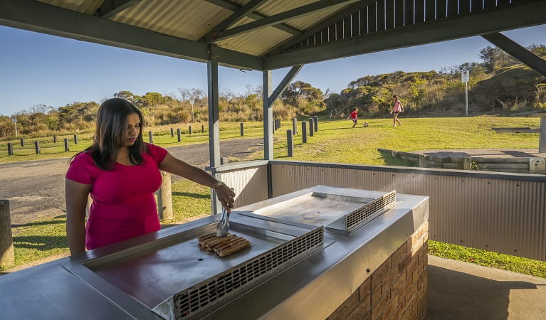 An adult cooking lunch on the barbecue and children playing soccer nearby, Frazer campground, Munmorah State Conservation Area. Photo: John Spencer/OEH