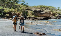 2 hikers walking across the black sand of Dark Beach, Murramarang National Park. Credit: Remy Brand &copy; Remy Brand