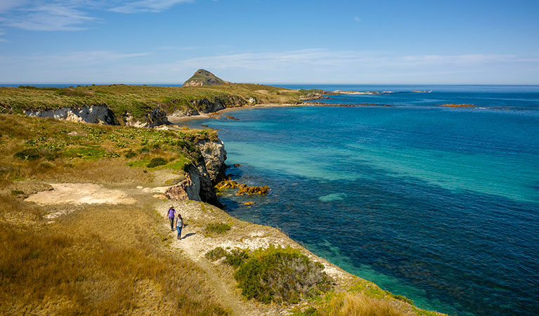 2 visitors walk along a path atop a coastal cliff, with the coastline and rocky headland in the background. John Spencer &copy; DPIE