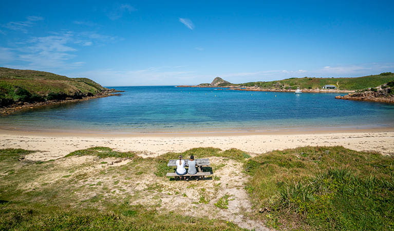 View of dunes, sandy beach and blue bay waters, with 2 people at a picnic table in the foreground.  John Spencer &copy; DPIE