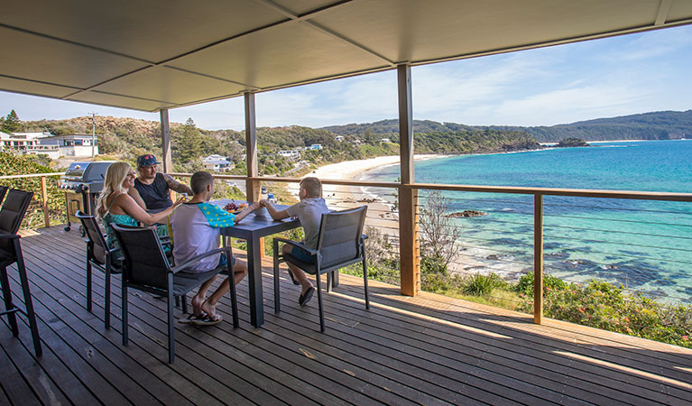 A family on the deck of Davies Cottage with Boat Beach and Sugarloaf Bay in the background. Photo: John Spencer &copy; DPIE