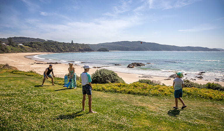 A family playing cricket on the grass below the cottage with Boat Beach in the background. Photo: John Spencer &copy; DPIE