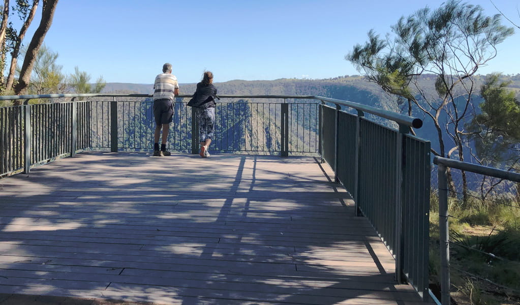 Two visitors stand on a large wooden viewing platform with metal railing and look out over a steep-sided valley. Photo &copy; Jessica Stokes