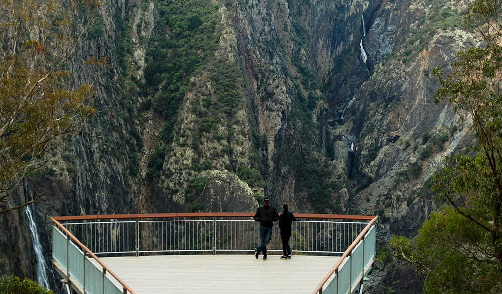 Two visitors stand on a large elevated viewing platform overlooking a steep valley with waterfalls. Photo credit: Leah Pippos &copy; DPIE