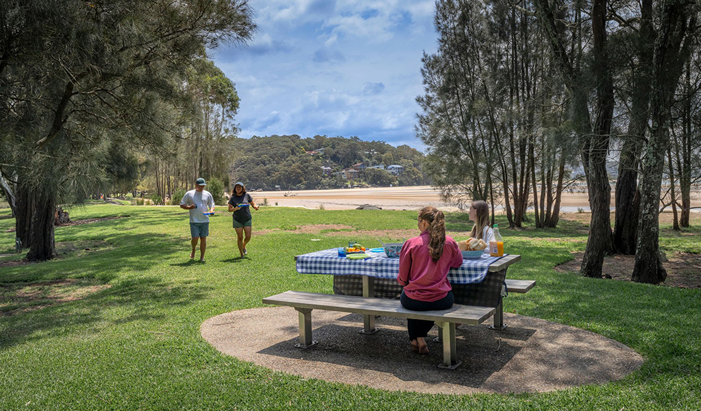 People having a picnic on a picnic table at Bonnie Vale campground next to the beach. Credit: John Spencer/DCCEEW  &copy; DCCEEW