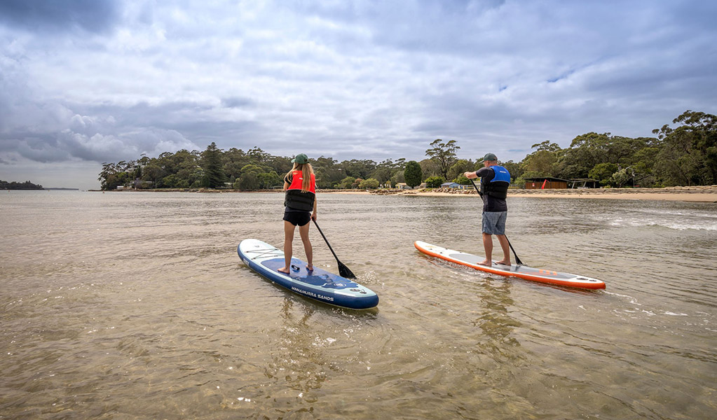 Two people paddle boarding on Hacking river next to Bonnie Vale campground. Credit: John Spencer © DCCEEW