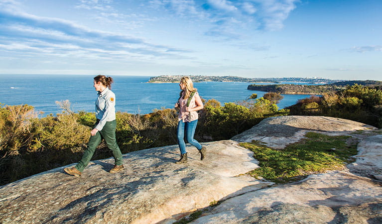 A field officer and park visitor walking along the rocks on Manly scenic walkway with a view of Middle Head in the background in Sydney Harbour National Park. Photo: Simone Cottrell &copy; OEH