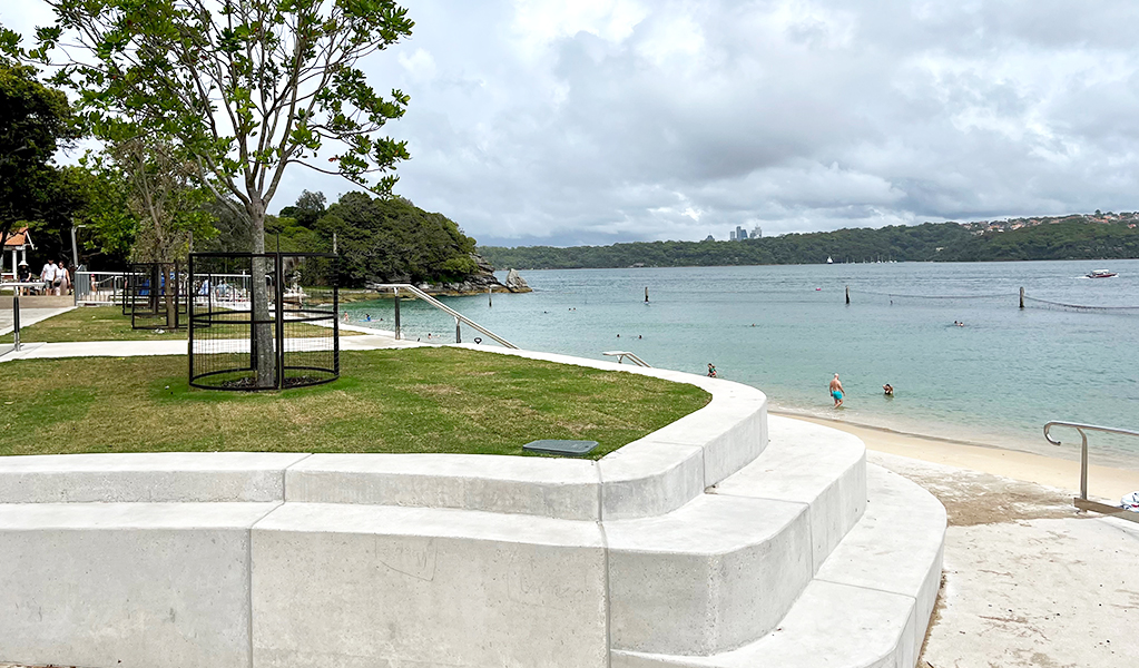 Curved concrete seawall with steps for seating with a grassy patch with trees at the top, and beach in the background. Photo: Natasha Webb &copy; DCCEEW