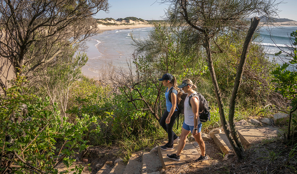 Two walkers approaching One Mile Beach with Samurai Beach in the background. Credit: John Spencer &copy; DPE