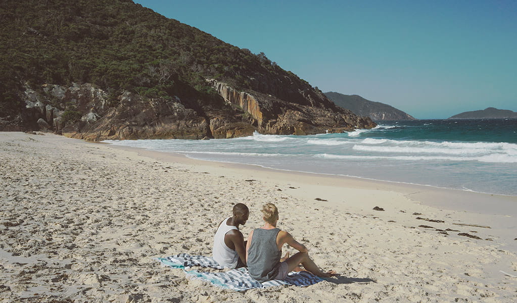 Two men sit on towels on the white sands of Box Beach, against a backdrop of breaking surf, blue ocean water and rugged rocky coastline. Photo: &copy; Erin McGauley