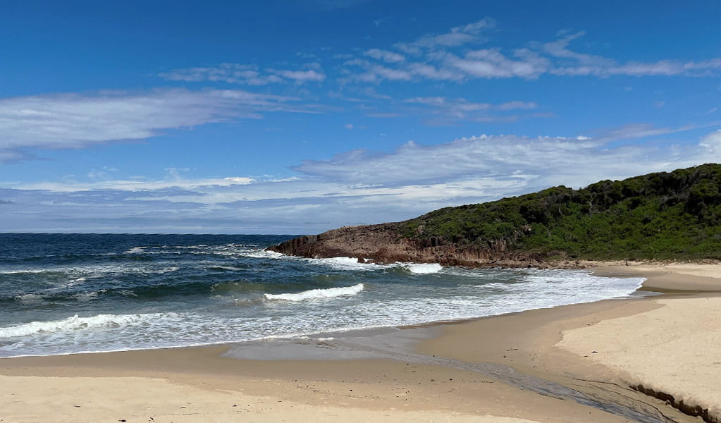 View of a sandy beach and breaking waves beneath a blue sky, with a rugged headland in the distance.  Photo: Josh Ford &copy; DPE