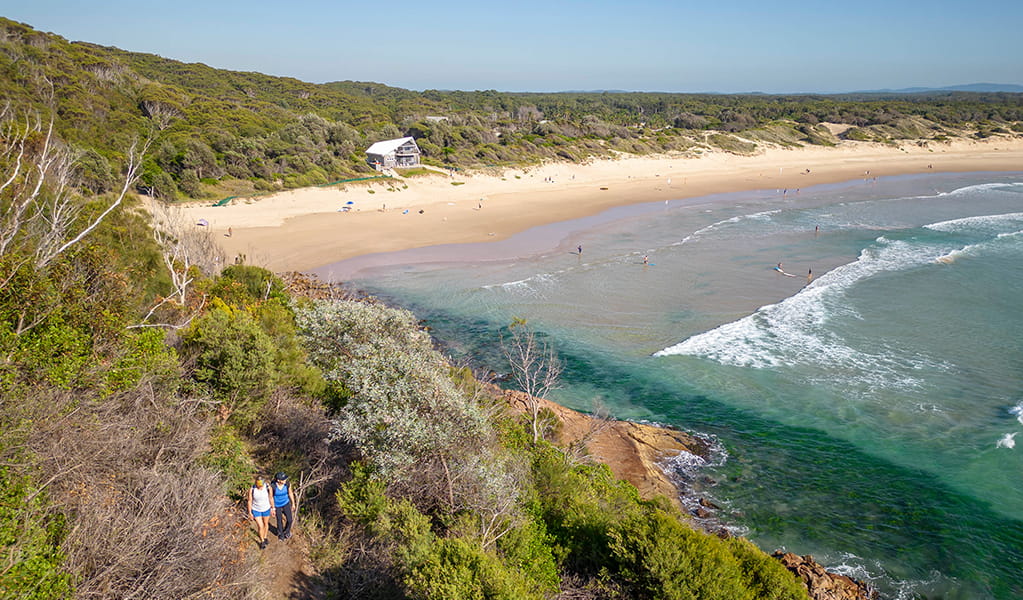 Aerial photo showing One Mile Beach, the surf life saving club, and walkers on a forested headland. Credit: John Spencer &copy; DPE