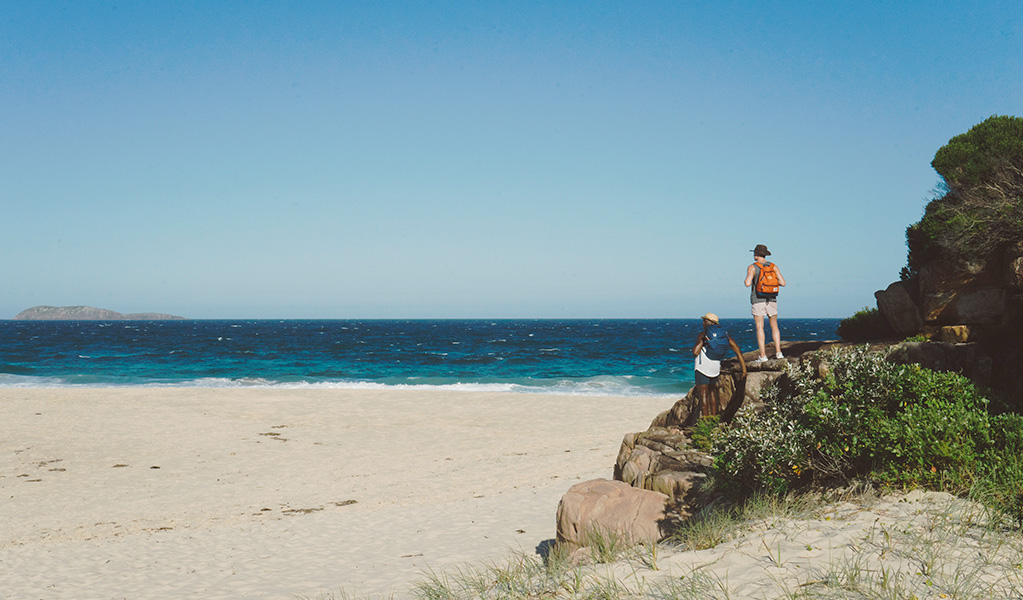 Two men wearing day packs stand near the base of a rocky headland, alongside the clean white sands and turquoise waters of Wreck Beach. Photo: &copy; Erin McGauley