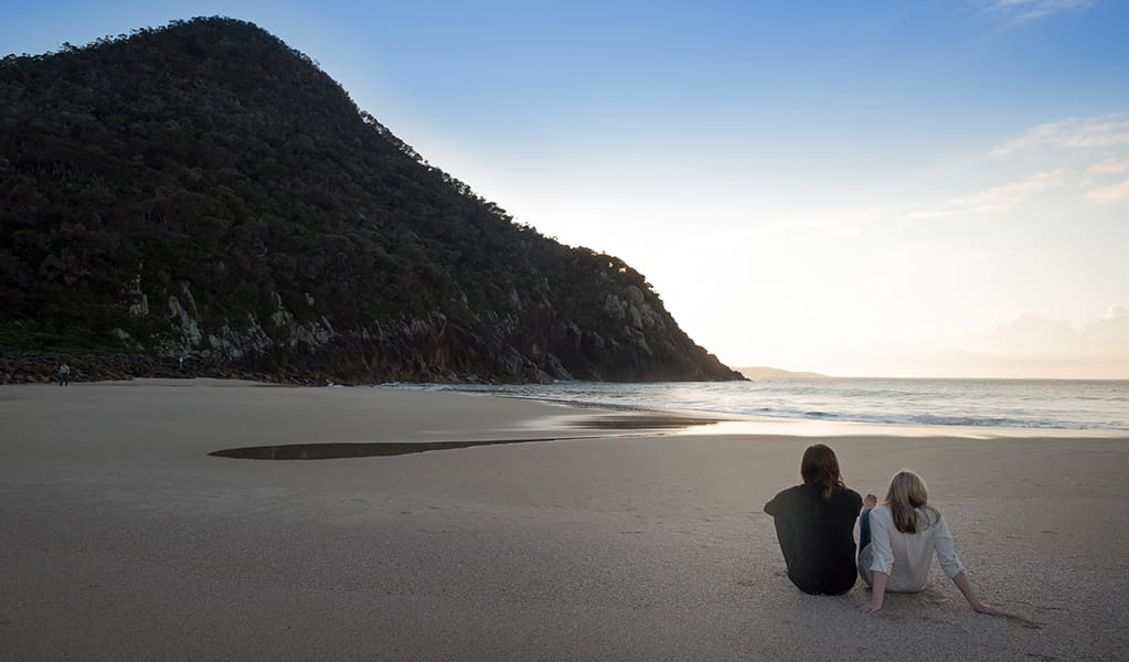 Couple seated on the sand of Zenith Beach at sunrise, with a dramatic rocky headland in the background.  Photo: John Spencer &copy; DPE