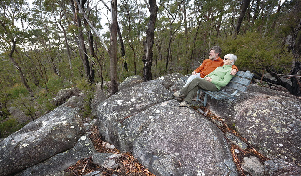 A couple dressed in warm clothes seated on a park bench atop a rock outcrop in a bushland setting. Photo credit: Rob Cleary &copy; DPIE