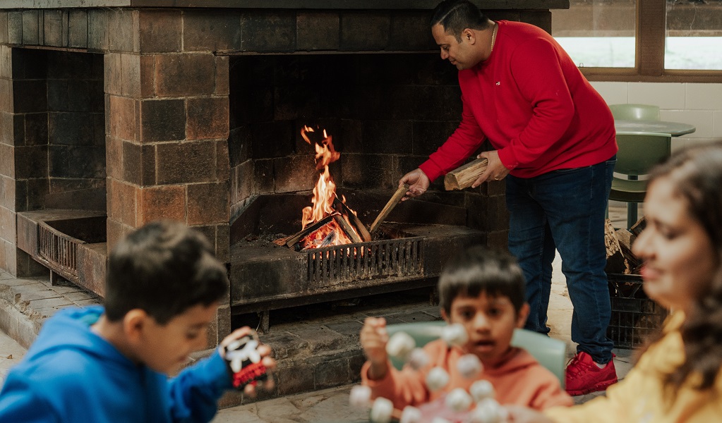 A man builds an open fire while his family prepare marshmallows for roasting, Barmah communal dining room. Credit: Remy Brand/DPE © Remy Brand