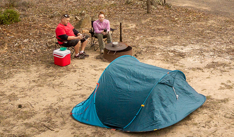 2 campers sitting next to a fire pit barbecue behind their tent. Photo: John Spencer/DPIE