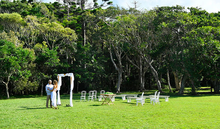 A couple standing next to a bridal arch with rainforest in the background at Broken Head lawn. Photo: Fiora Sacco &copy; DPIE