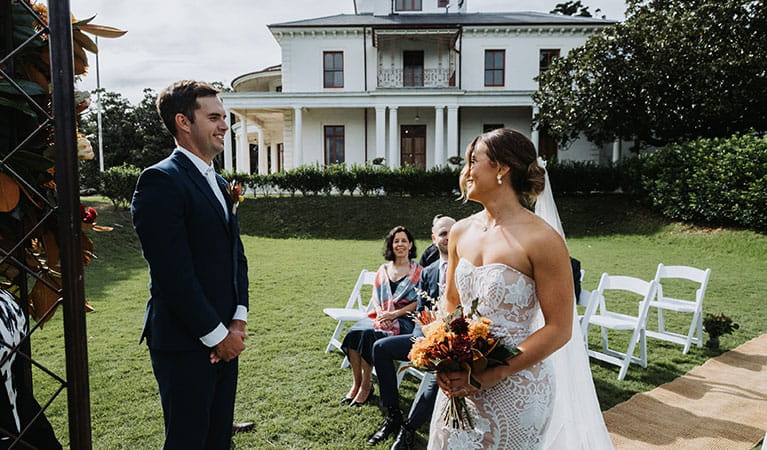 A bride and groom with seated guests and Strickland House in the background at Strickland Estate in Sydney Harbour National Park. Photo &copy; Matt Horspool