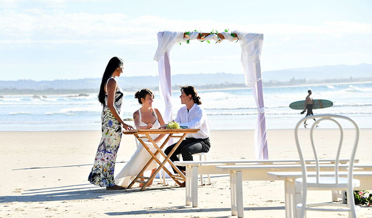 A couple and their celebrant at at a signing table with a wedding arch and ocean in the background on Wategos Beach. Photo: FIora Sacco &copy; DPIE