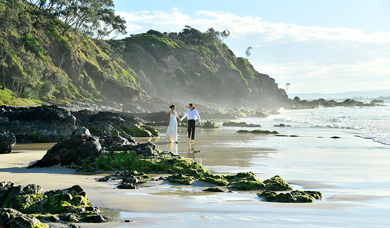 A couple walking past rocks and tidal pools on Wategos Beach in Cape Byron State Conservation Area. Photo: Fiora Sacco &copy; DPIE