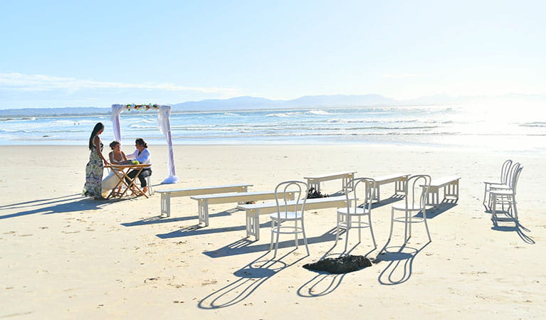 A couple and their celebrant at a signing table with chairs and benches in the foreground on Wategos Beach. Photo: Fiora Sacco &copy; DPIE