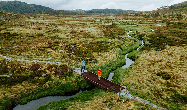 2 hikers crossing a creek between Charlotte Pass Village and Perisher Valley in Kosciuszko National Park. Credit: Daniel Parsons / DCCEEW © Caravel Content